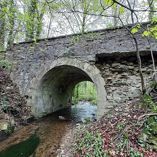 Seitliche Ansicht einer Brücke aus Stein, die über einen Bach führt. Es sind beschädigte Bereiche zu erkennen.