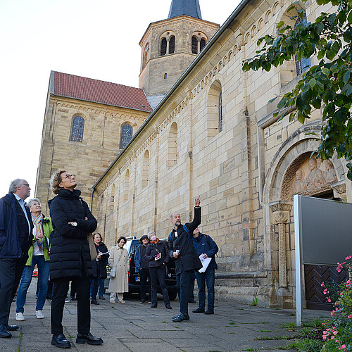 Ein Mitarbeiter der Klosterkammer schildert einer großen Gruppe während einer Tagung die Geschichte der Kirche Sankt Godehard in Hildesheim. Sie stehen vor der Fassade.