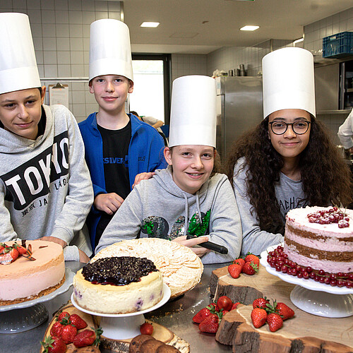 Vier Jugendliche mit Kochmützen stehen in einer professionellen Küche vor einem Tisch mit Torten und Kuchen.