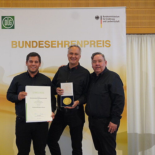 Drei schwarz gekleidete Männer mit einer Urkunde und einer Medaille stehen vor einem Roll-up.