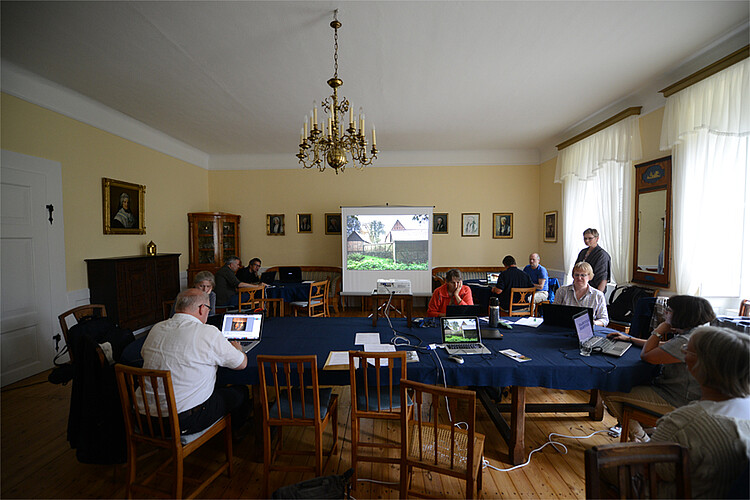 Mehrere Personen sitzen vor Laptops an Tischen in einem Saal. Hinten steht eine Leinwand, auf die ein Foto projiziert ist.