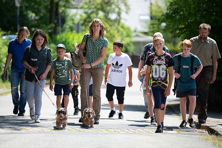 Eine Gruppe von Schulkindern läuft von einer Försterin und einem Förster der Klosterforsten auf einer Straße in ein Waldstück.