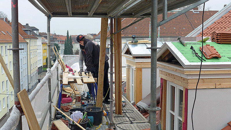 Zwei Handwerker stehen oben auf einem Gerüst und arbeiten an Holzteilen für Dachgauben.