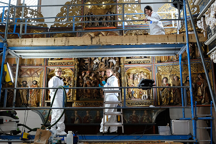 Auf einem zweistöckigen Gerüst vor einem reichlich verzierten Altaraufsatz arbeiten drei Frauen in Schutzanzügen.