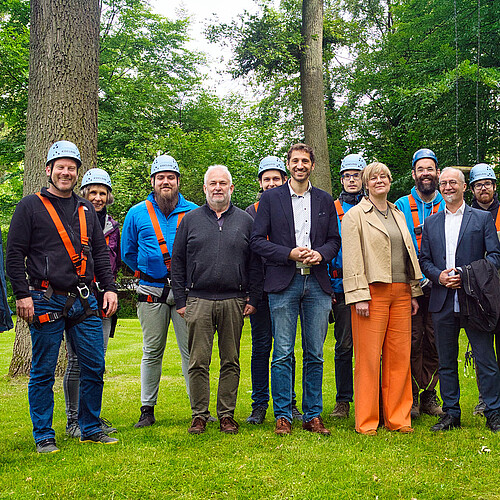 Gruppenbild mit 13 Personen vor hohen Bäumen. Einige tragen Helm und Klettersteiggurt.