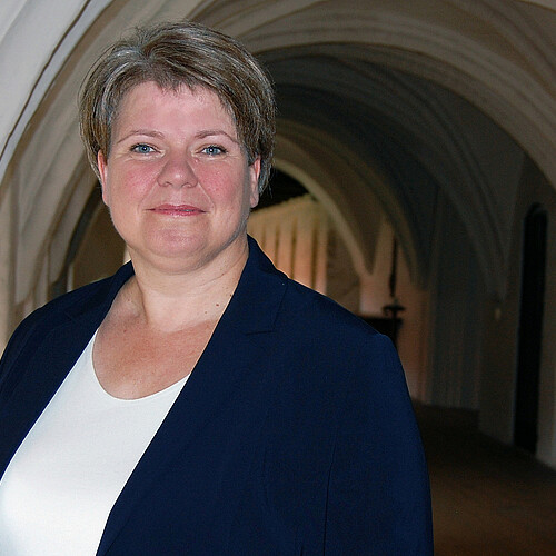 Portrait einer Frau vor Kirchengewölbe, sie trägt einen blauen Blazer und kurze Haare.