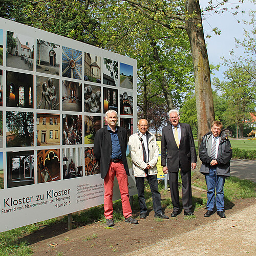 Drei Personen stehen vor einer drei mal vier Meter großen Tafel, auf der 28 quadratische Fotos zu sehen sind.