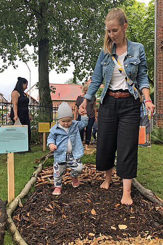 Eine junge Frau geht barfuß mit einem Kleinkind an der Hand über einen Barfußpfad mit unterschiedlichen Materialien aus dem Wald.