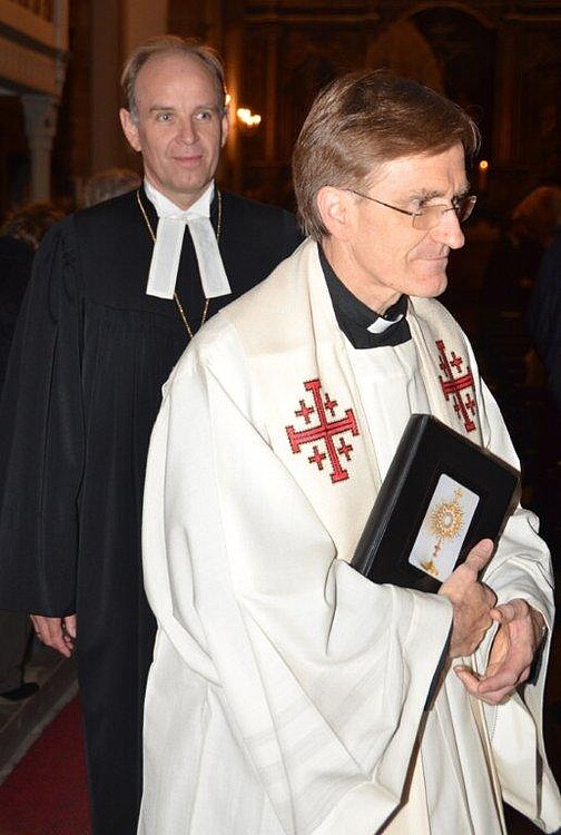 Zwei Herren in geistlichen Gewändern gehen hintereinander den Gang einer Kirche entlang.