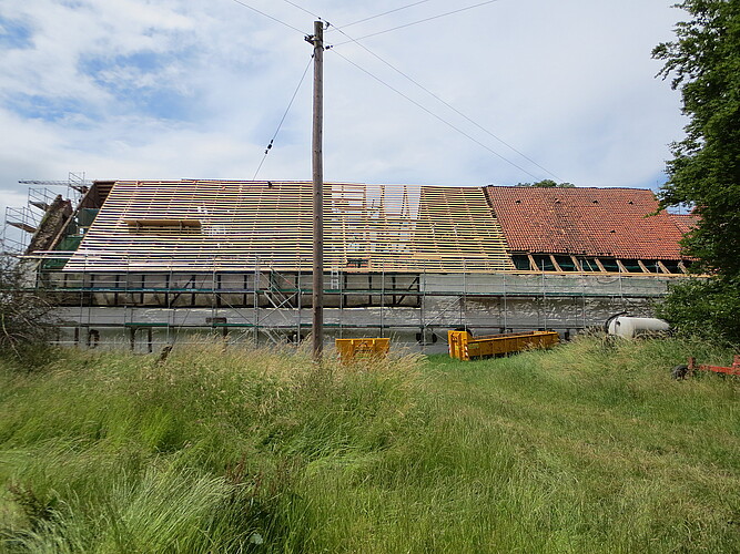 Ein langes Gebäude von der Längsseite - das Dach ist abgedeckt, neue Sparren sind zu sehen.