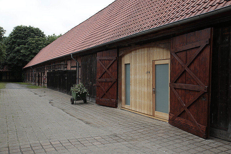 Eine langgezogene Scheune ist mit roten Dachziegeln gedeckt - ein Tor steht offen.