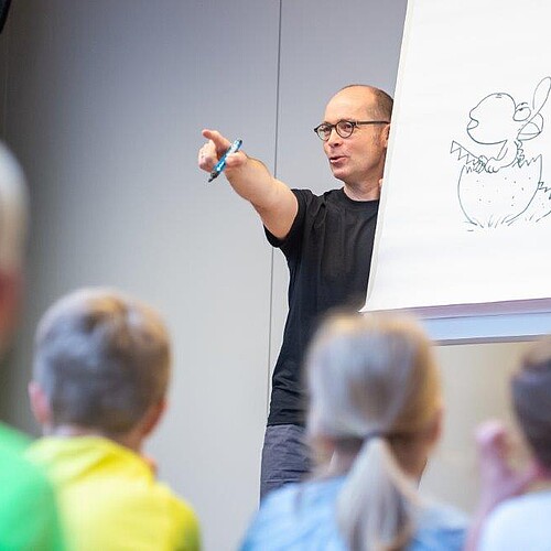 Ein Mann mit einem Stift in der Hand steht neben einem Flipchart mit einer Zeichnung, davor sitzen Zuschauer.