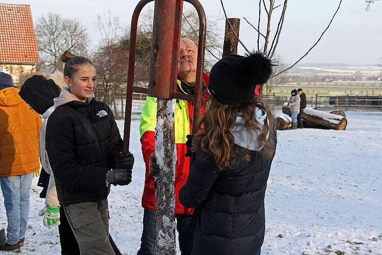 Zwei Schülerinnen in Winterbekleidung verwenden eine Pfahlramme aus Metall, um einen Pfahl zur Befestigung eines Jungbaums in die Erde zu rammen.