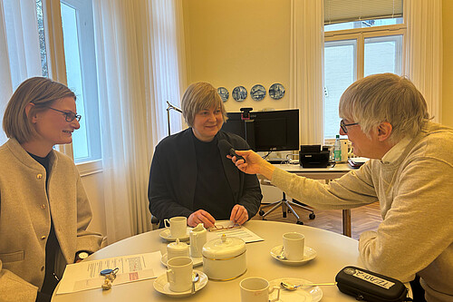 Ein Mann hält ein Mikrofon, eine Frau spricht hinein. Eine weitere Frau sitzt mit am Tisch.