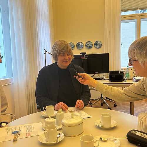 Ein Mann hält ein Mikrofon, eine Frau spricht hinein. Eine weitere Frau sitzt mit am Tisch.