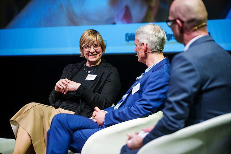 Eine Frau und zwei Männer sitzen auf einer Bühne, die Frau spricht.