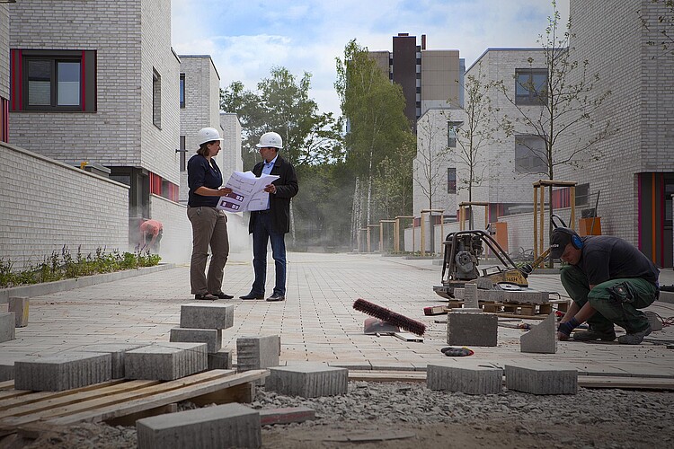 Zwei Personen mit Schutzhelmen stehen auf einer Straße in dem Neubaugebiet Lehnbachhöfe, um sie herum wird gebaut.