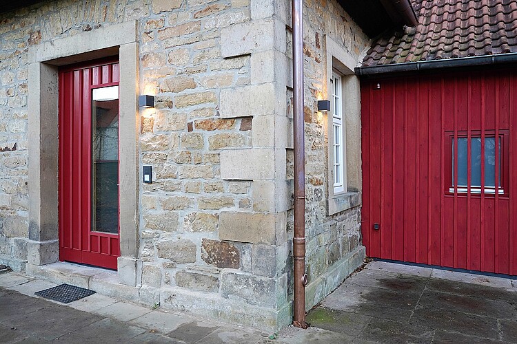 Eine Außenwand aus hellem Stein mit einer roten Eingangstür aus Holz.