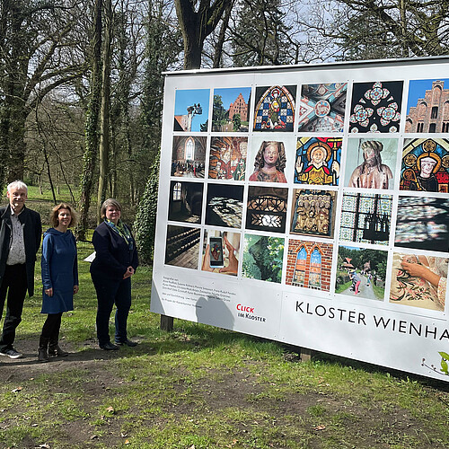 Drei Personen stehen neben einer großformatigen Tafel mit 28 quadratischen Fotos.