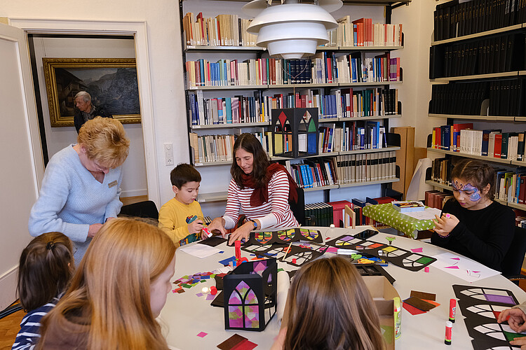 Um einen großen runden Tisch in der Bibliothek der Klosterkammer haben sich zwei Frauen und einige Kinder versammelt, die gemeinsam aus buntem Papier Laternen basteln.