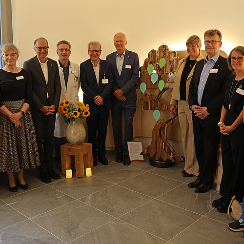 Zehn Personen stehen in einem Halbkreis, in der Mitte befindet sich ein großer Holzbaum.