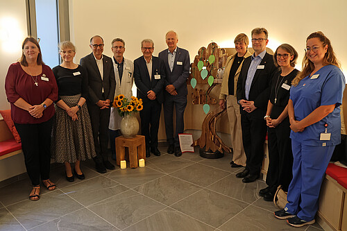 Zehn Personen stehen in einem Halbkreis, in der Mitte befindet sich ein großer Holzbaum.