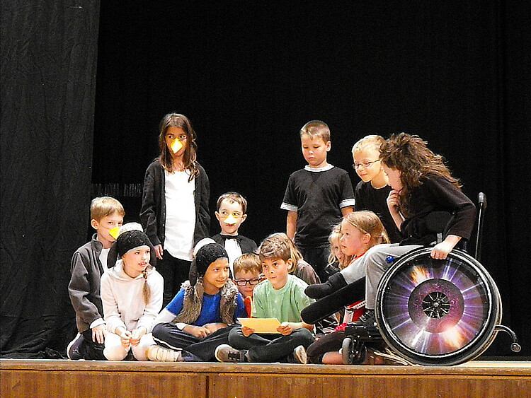 Grundschulkinder stehen und sitzen zusammen auf einer Bühne - darunter ist ein Mädchen im Rollstuhl.