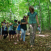 Eine Reihe von Kindern mit zugebundenen Augen, die sich jeweils an den Schultern fassen, läuft unter der Leitung von Försterin Stefanie Grevelhörster durch den Wald.