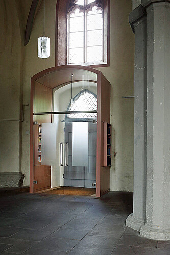 Der Eingangsbereich zu einer Kirche von innen fotografiert. Um die Tür herum ist ein Kasten gebaut, der mit einer Glastür abschließt.