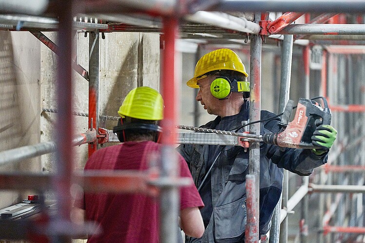 Zwei Arbeiter mit Bauhelm und Gehörschutz stehen auf einem Gerüst; einer von ihnen bohrt mit eine großen Bohrmaschine in Stein.