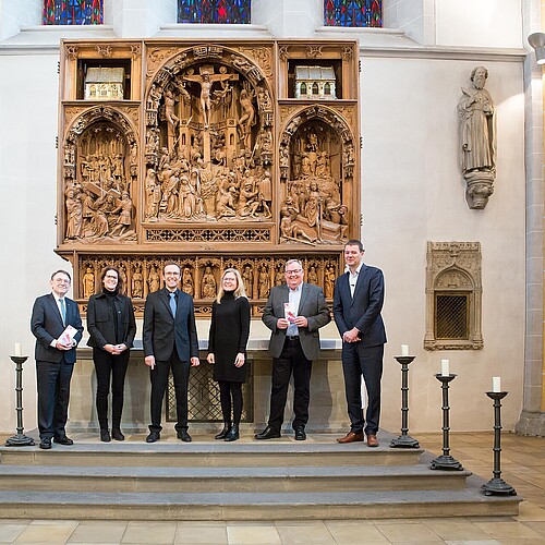 Sechs Personen in dunkler Kleidung stehen in einer Kirche vor einem hölzernen Altaraufsatz.