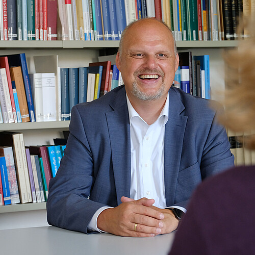 Ein Mann in blauen Jackett und weißen Hemd sitzt vor Bücherregalen.