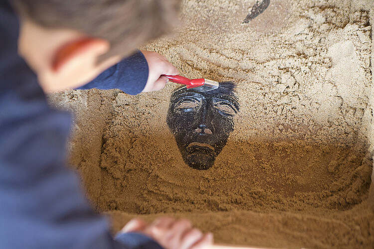 Ein Junge legt vorsichtig eine Maske aus Metall unter Sand frei.