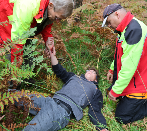 Auf Waldboden liegt bei einer Übung eine scheinbar verletzte Person, zwei Helfende links und rechts daneben