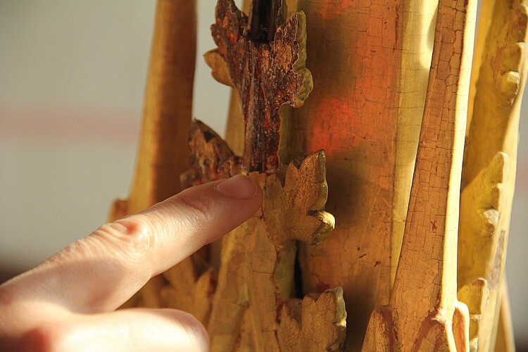Ein Stück der hölzernen Verzierung von der die goldene Farboberfläche abgesprungen ist, ein Finger zeigt darauf.